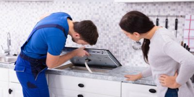 Woman Looking At Serviceman In Uniform Fixing Induction Stove In The Kitchen